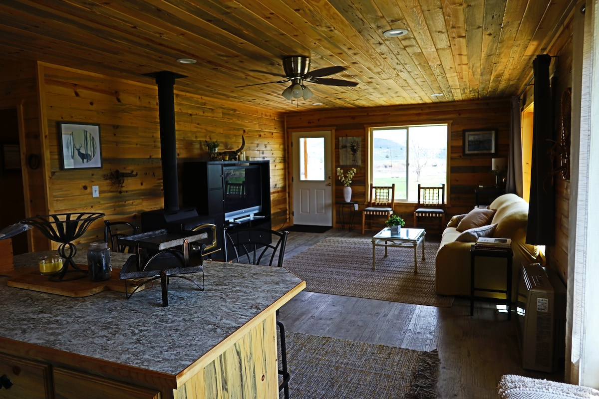The living area features natural wood paneling and a ceiling fan. A cozy seating arrangement includes a sofa and two chairs facing a large window. An open kitchen space with rustic countertops and appliances is visible, while a warm, inviting ambiance is created by natural light.