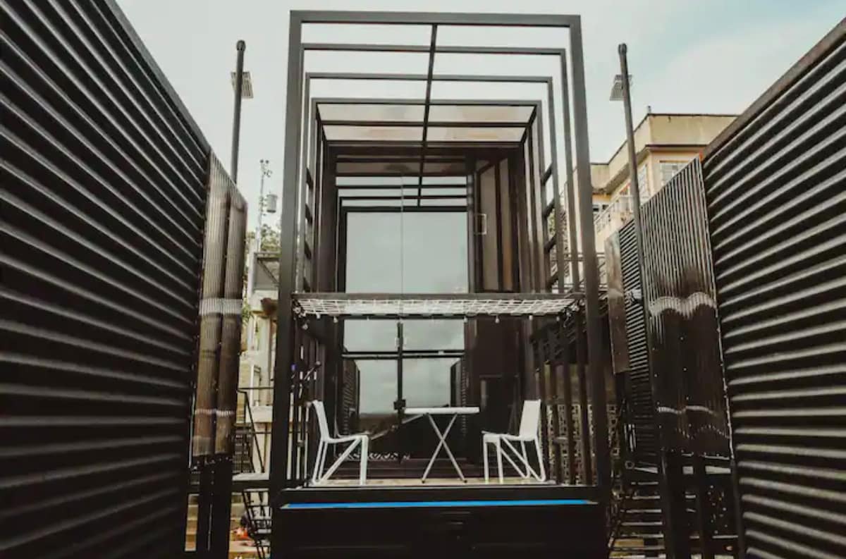 A sleek, modern tiny house is framed with black metal structures, featuring a front deck. Positioned on the deck are a small table and two white chairs, creating a cozy outdoor seating area. Large glass windows allow natural light to fill the interior.