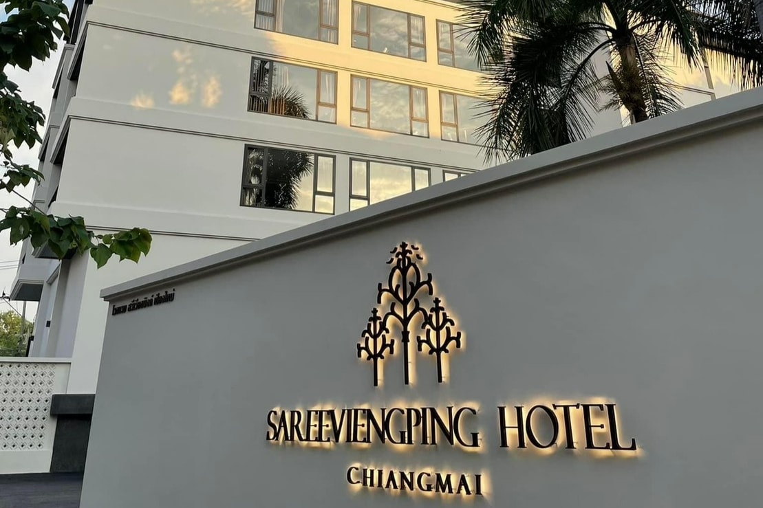 The exterior of the SAREEVIENGPING Hotel in Chiang Mai is showcased, featuring a modern design with a light-colored facade. The hotel name is prominently displayed on a stylish sign, illuminated against the building, surrounded by greenery.