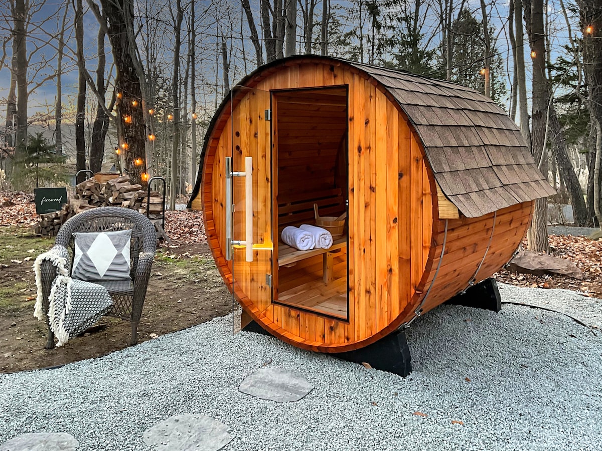 A private outdoor steam sauna constructed from natural wood features a rounded barrel design. The entrance to the sauna is highlighted by a glass door, with neatly arranged towels placed inside. Nearby, a cozy wicker chair and decorative cushions are positioned on a gravel surface, surrounded by trees.