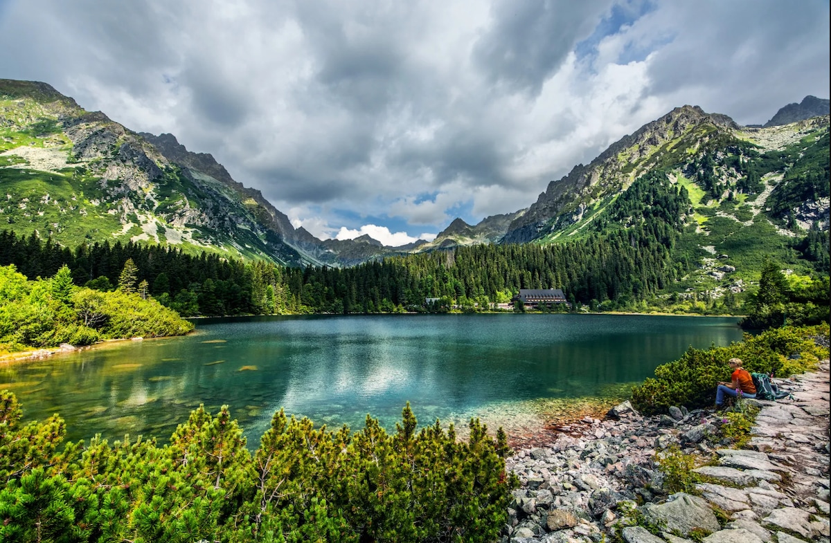 A serene lake is framed by lush green trees and dramatic mountain peaks under a partly cloudy sky. The calm water reflects the surrounding landscape, while a rocky shoreline provides texture in the foreground. A small figure can be seen peacefully seated by the water's edge.
