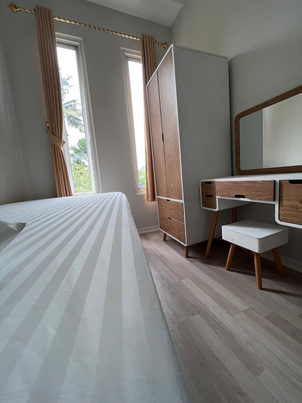 The minimalistic bedroom features a spacious bed with striped linens, paired with a sleek wooden wardrobe and a dressing table. Natural light enters through tall windows adorned with drapes, illuminating the soft color palette of the room.