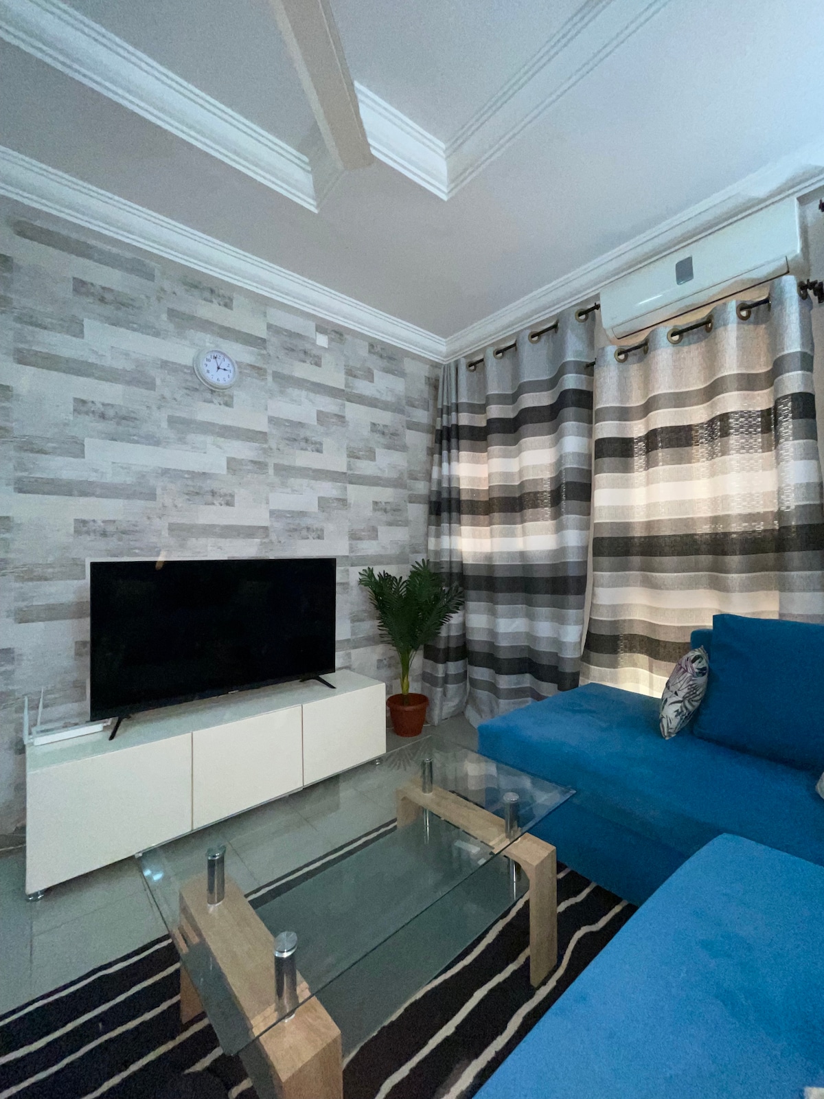 A modern living room features a large flat-screen TV set on a white media console. A blue sectional sofa is positioned against a textured wall adorned with neutral tones. Vertical striped curtains frame the window, and a potted plant adds a touch of greenery.