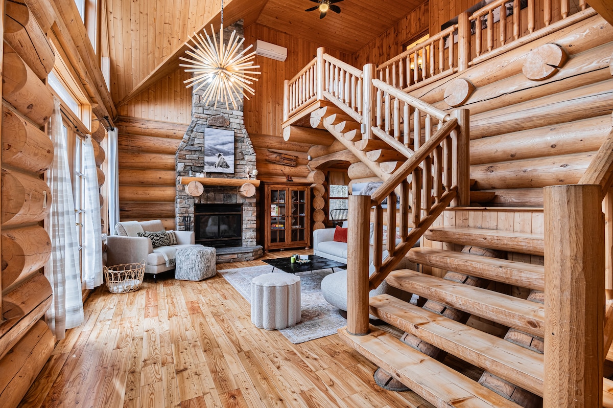 A spacious living area features a stone fireplace as a focal point, surrounded by comfortable seating. Natural wood finishes create warmth, while the wooden staircase adds character. Large windows allow ample light, enhancing the inviting atmosphere of the chalet.