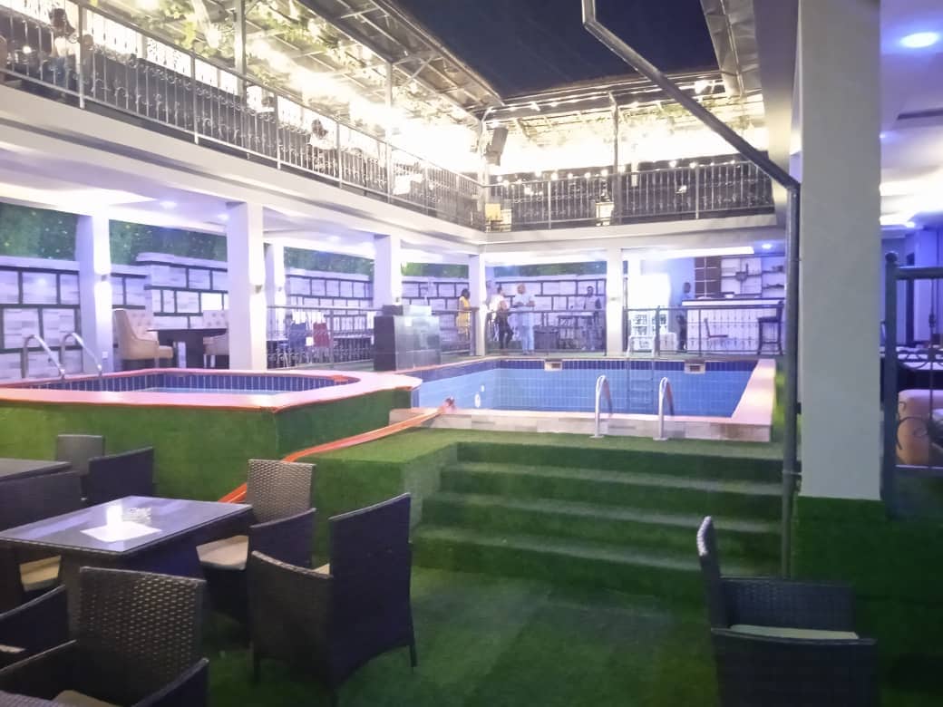 An outdoor area is presented with an inviting swimming pool surrounded by green grass. Lounge chairs are arranged nearby, while covered seating is visible at a lower level. The space is illuminated with soft lighting, creating a serene atmosphere in the evening.