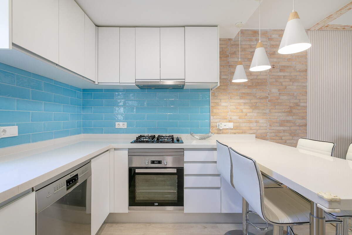 A modern kitchen features a sleek design with white cabinetry and a vibrant blue backsplash. Stainless steel appliances, including a gas stove and an oven, are complemented by a spacious countertop with three white bar stools. Pendant lights hang above, enhancing the kitchen's contemporary feel.
