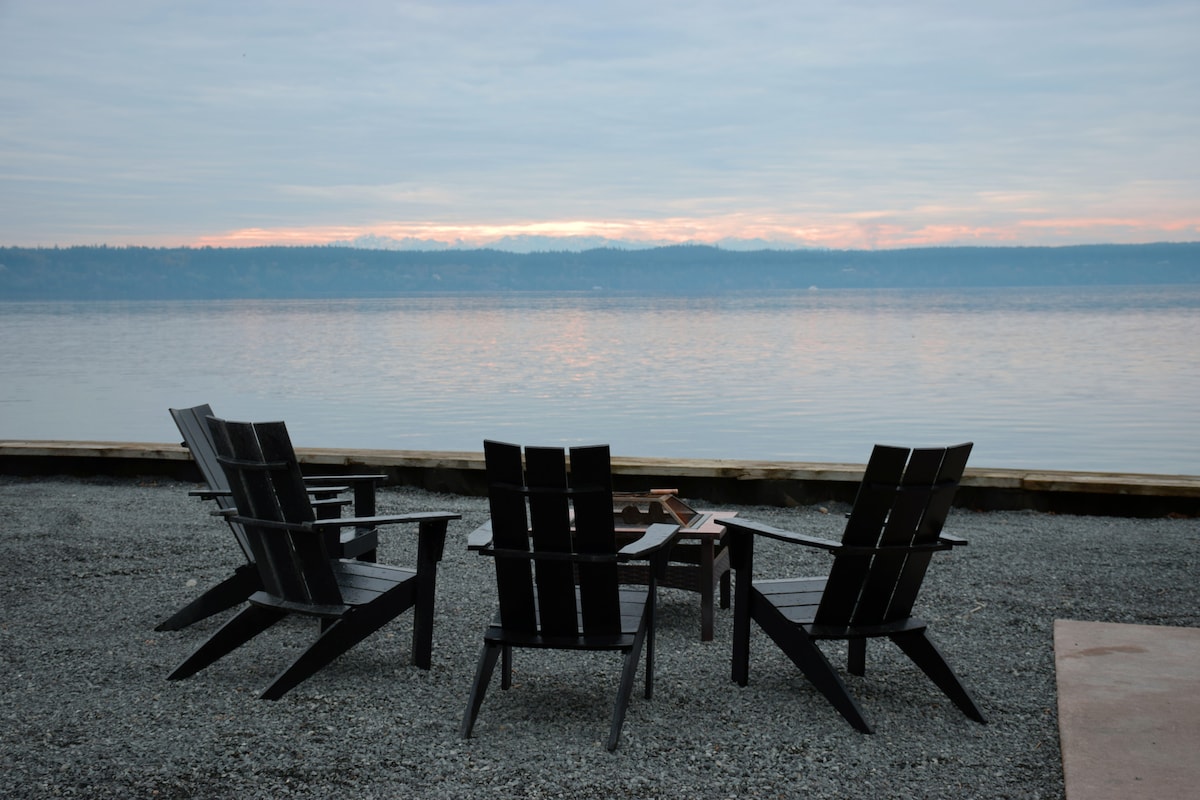 A tranquil outdoor seating area features four Adirondack chairs arranged around a fire pit. The gravel surface offers a natural setting, with water views reflecting soft hues of early morning light. The surrounding landscape showcases serene waterfront and distant hills.