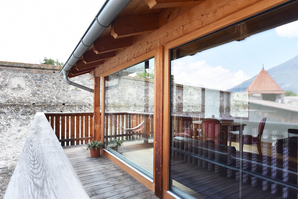 An outdoor balcony is framed by wooden beams, featuring panoramic glass walls that provide views of the surroundings. A pot with greenery is placed by the railing, and a cozy seating area is visible, inviting guests to enjoy the fresh air in a peaceful setting.