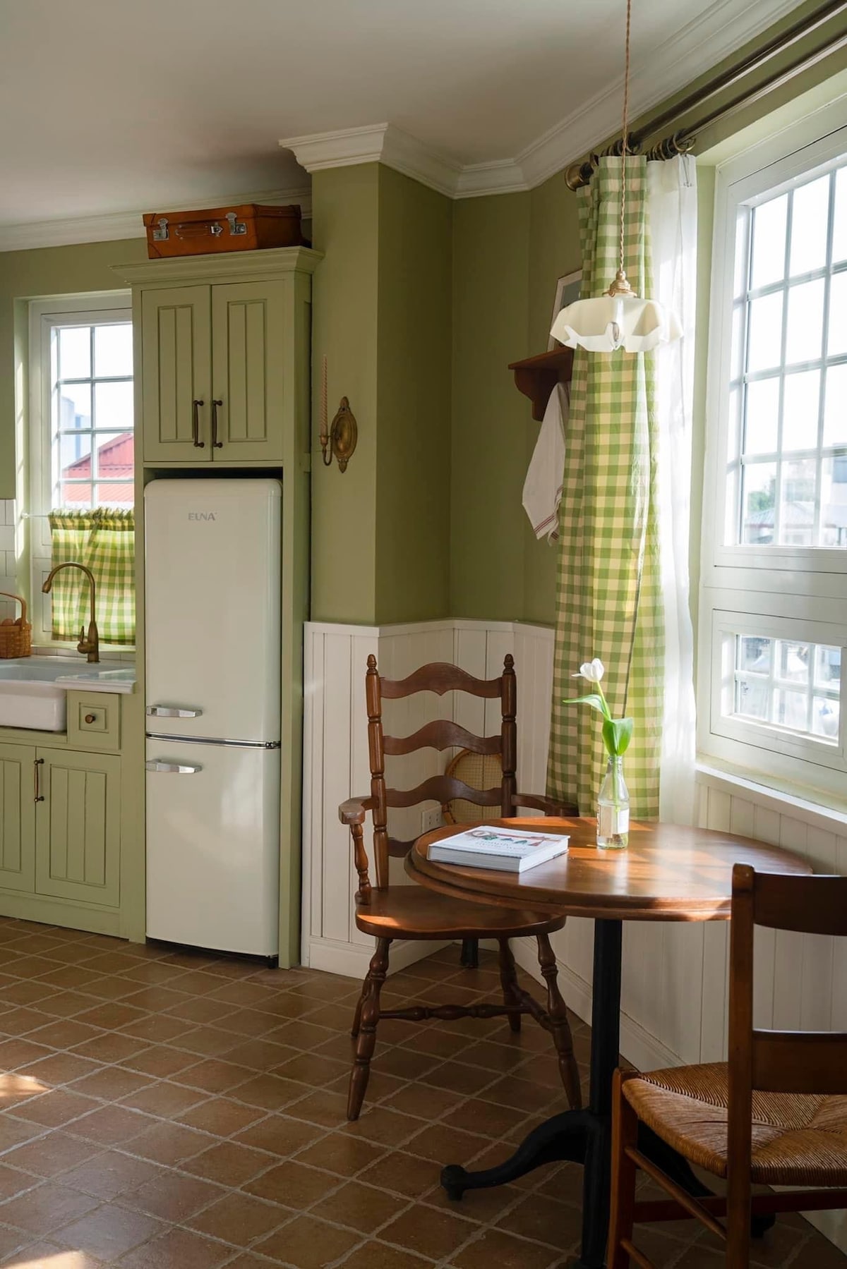A sunlit kitchen features light green cabinetry and a white refrigerator, with natural light filtering through window panes. A small round table is paired with wooden chairs, and a vase with a single tulip adds a touch of freshness to the space.