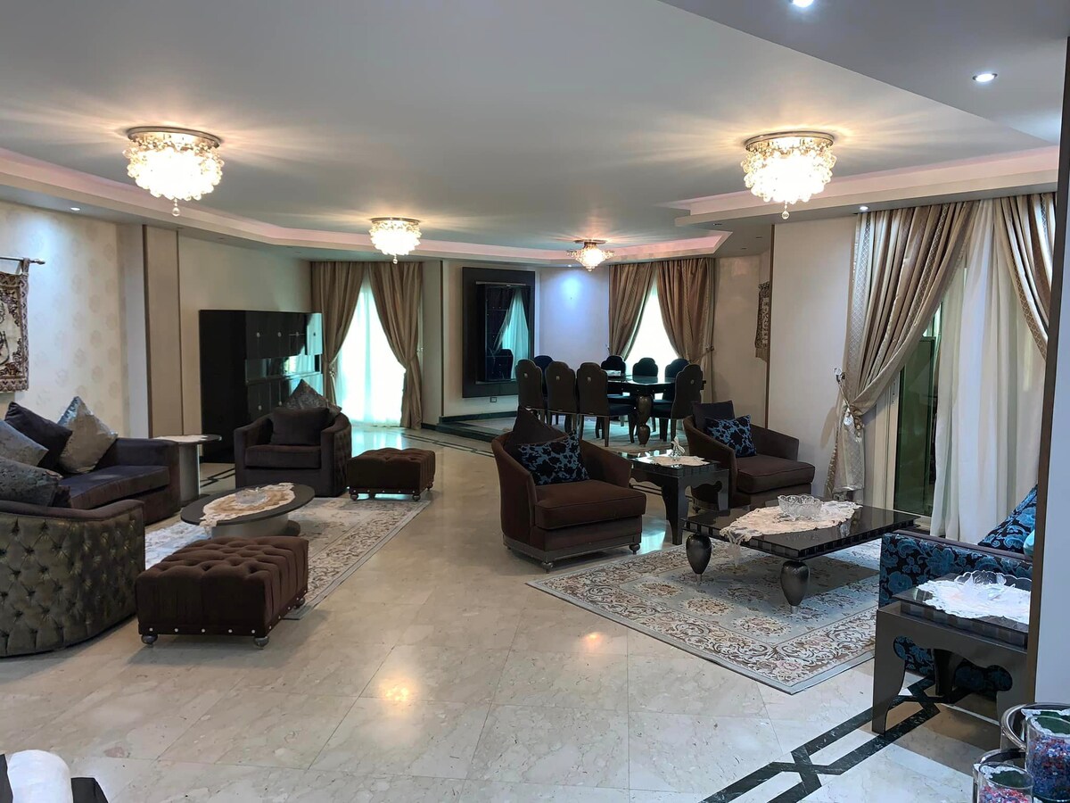 A spacious living area features elegant seating arrangements with plush chairs and sofas. Decorative chandeliers illuminate the room, while large windows allow natural light to fill the space. A large dining table is situated nearby, enhancing the family-friendly ambiance.