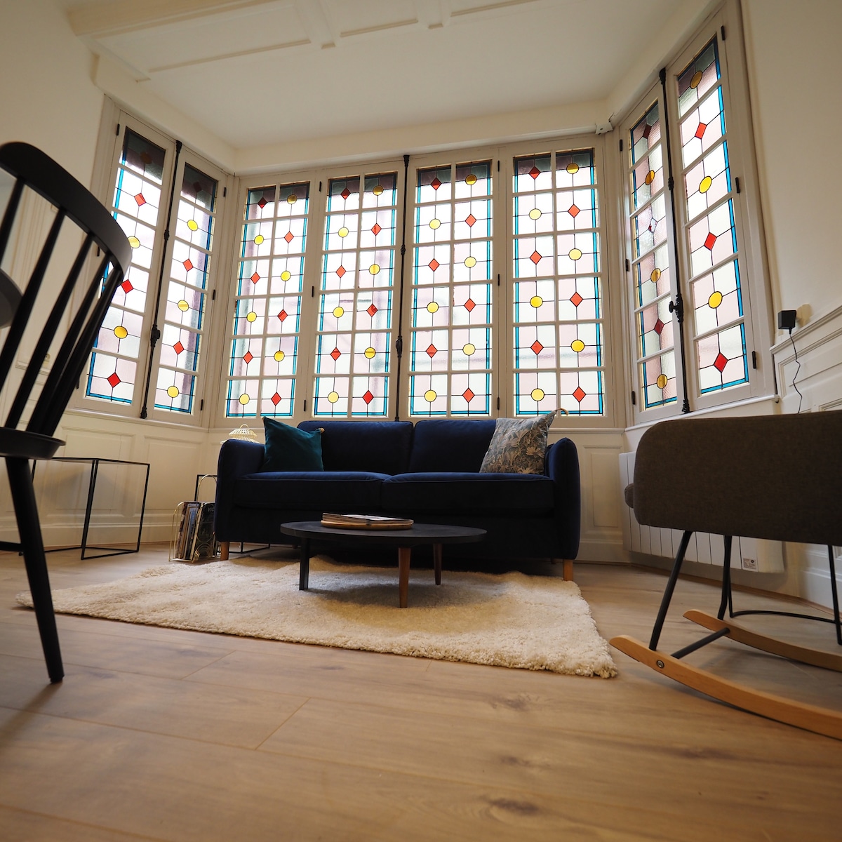 The living area is adorned with a bow window featuring vibrant stained glass, which floods the space with natural light. A dark blue sofa is positioned in front of a low coffee table on a soft area rug. A rocking chair and a small side table are also included in the cozy setup.