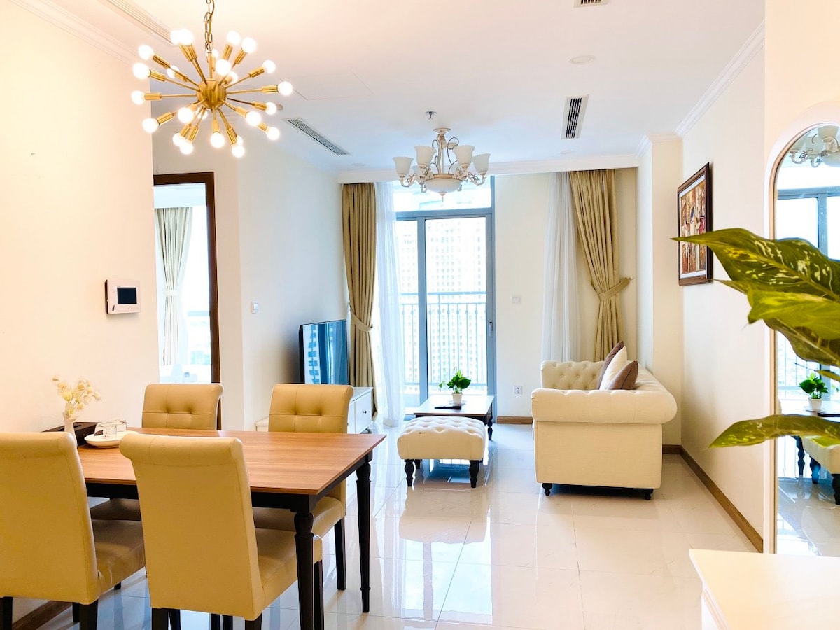 A bright living area is presented with large windows allowing natural light to fill the space. A light-colored sofa complements a wooden dining table surrounded by upholstered chairs. A decorative chandelier hangs above, enhancing the room's modern design.