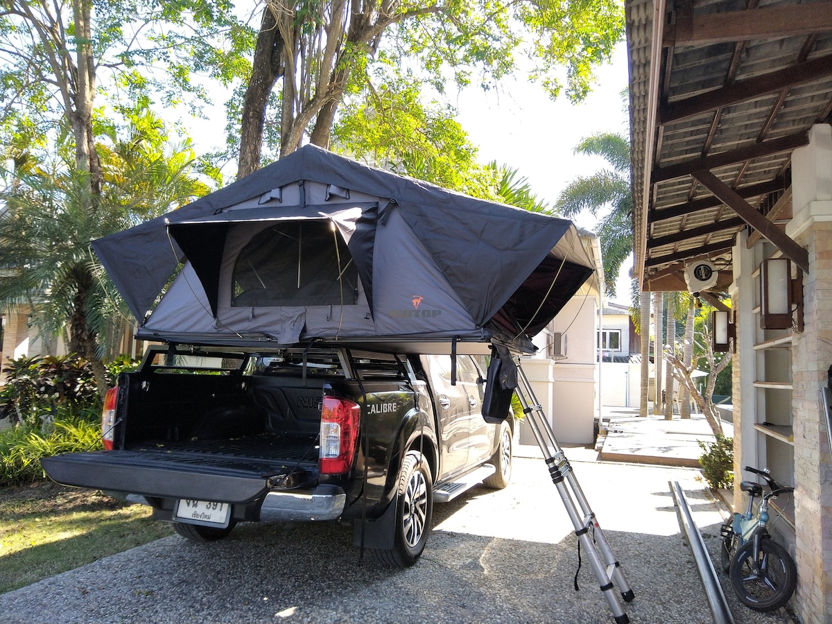 A vehicle with a rooftop tent is parked on a gravel drive, showcasing the tent's open flaps for ventilation. Surrounding greenery and palm trees provide a natural backdrop, while a building with a sloped roof is partially visible on the side.