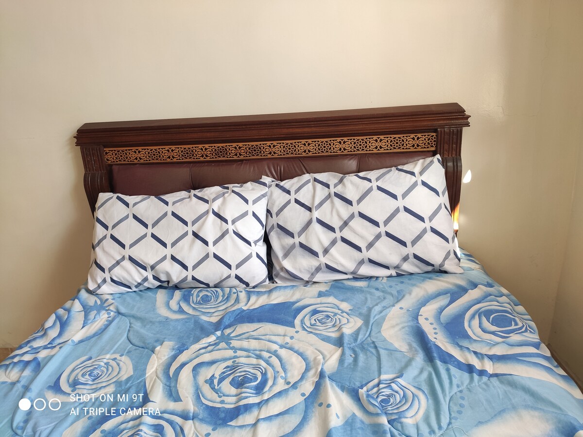 A bed with a decorative wooden headboard is centered against a neutral wall. Two patterned pillows rest against the headboard, while a soft blue and white floral blanket covers the bed, creating a calming visual appeal in the room.
