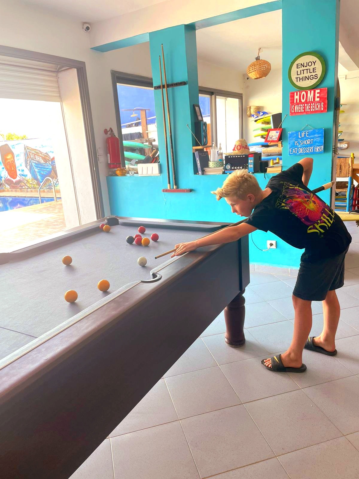 A game room is depicted, featuring a well-maintained billiard table positioned under natural light from large windows. Colorful decor enhances the inviting space, with a collection of vibrant surfboards visible in the background. The floor is tiled, complementing the overall bright atmosphere.