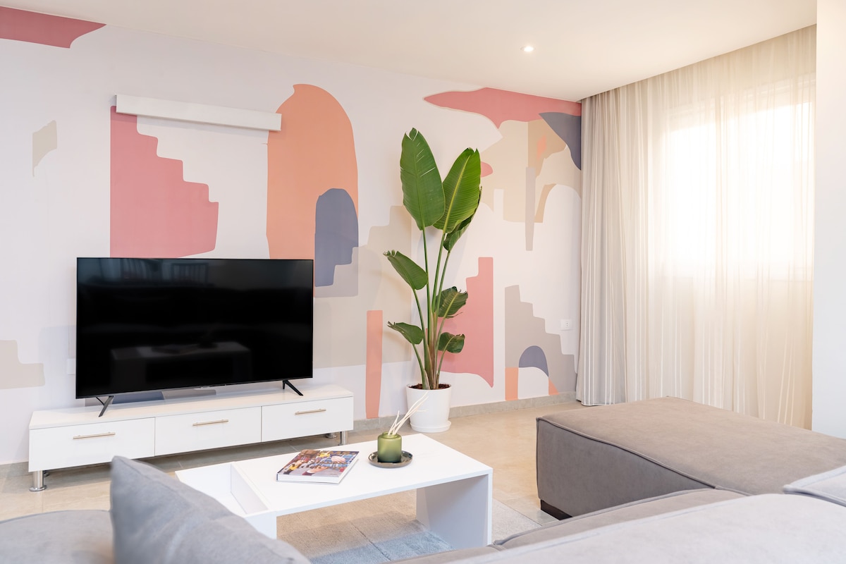 A welcoming living area features a large smart TV placed on a white media unit. A potted plant adds a touch of greenery. Light-colored, soft furnishings create a cozy atmosphere, while a colorful mural adorns the wall, contributing to the stylish decor.