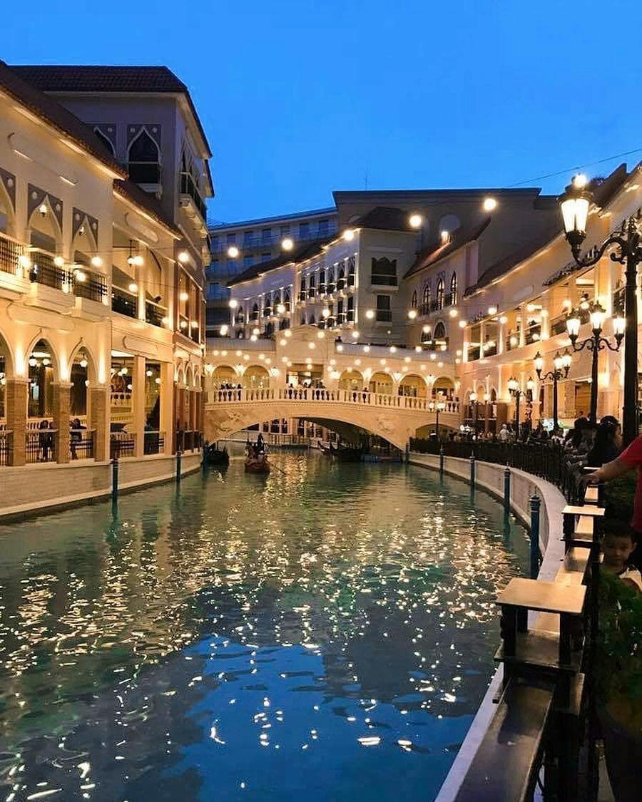 An evening view of a vibrant canal area is presented, featuring charming European-style architecture. The scene showcases warm lighting along the buildings and bridges, reflecting on the water's surface, enhancing the inviting ambiance of this lively tourist destination.