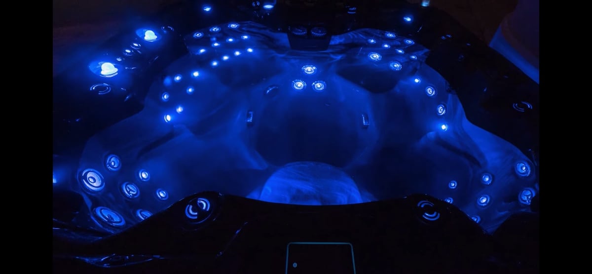 A spacious hot tub is illuminated with soothing blue lights, featuring multiple jets arranged around its interior. The sleek black exterior showcases buttons for easy control, while the inviting water surface reflects the ambient glow, providing a relaxing atmosphere for unwinding.