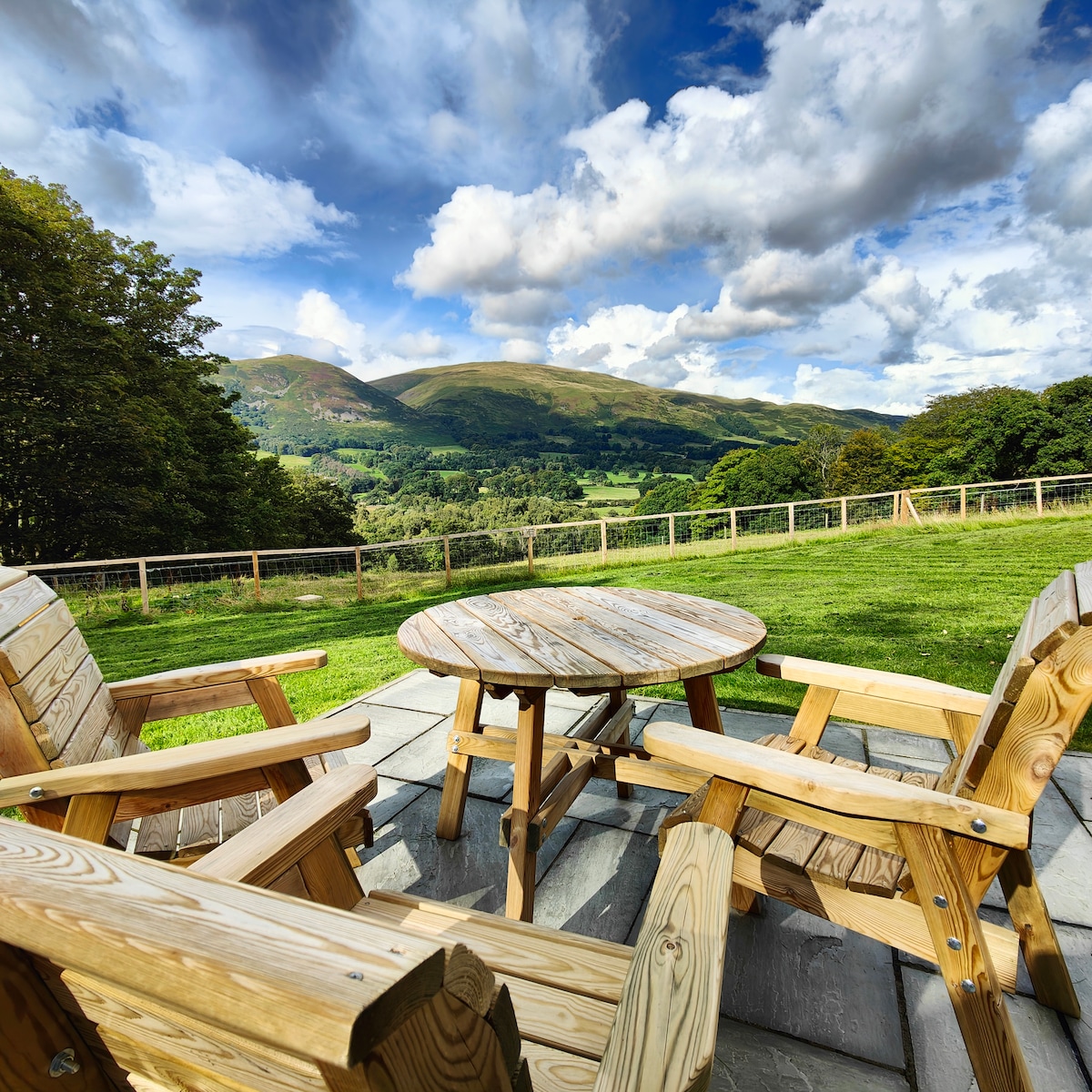 A wooden table surrounded by two matching chairs is positioned on a stone patio. The scenic view features rolling hills under a partly cloudy sky, with a green landscape extending to the horizon. A wooden fence outlines the area, enhancing the tranquil setting.
