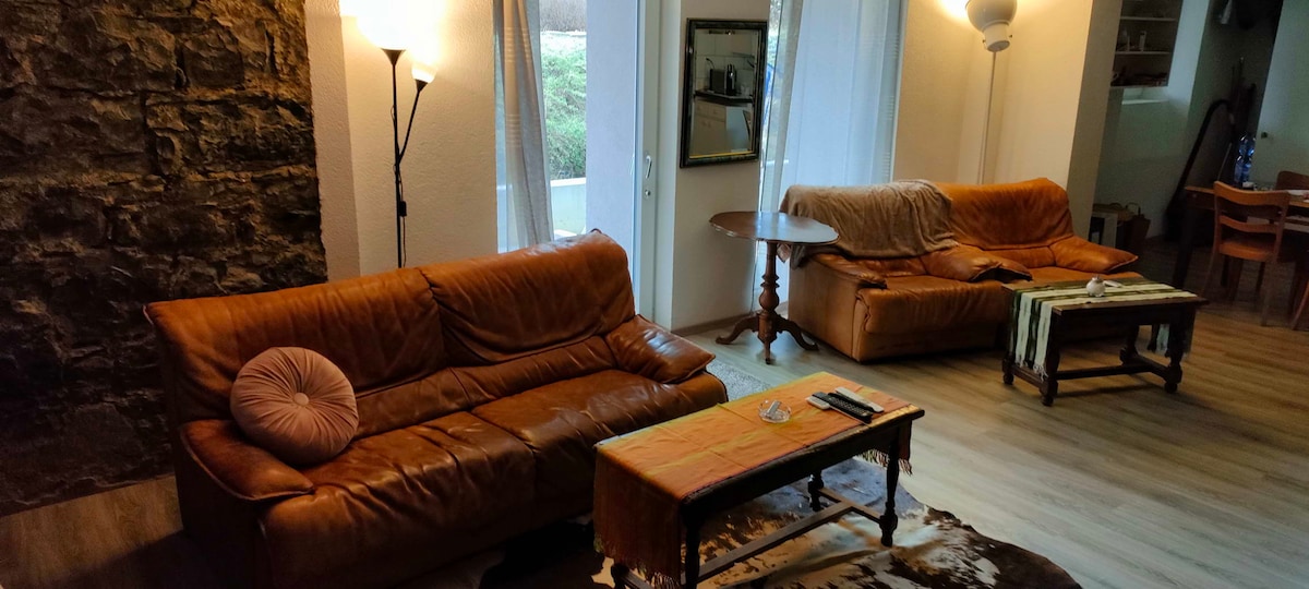 A living area showcases two brown leather sofas positioned around a wooden coffee table. Natural light emits from large windows, highlighting the textured stone wall in the background. A mirror and decorative items add character to the warm space.