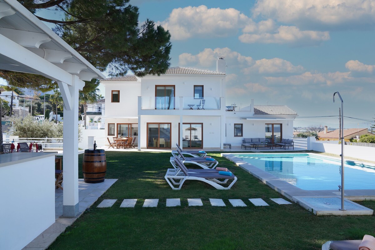 The villa is surrounded by a neatly maintained lawn, with a swimming pool inviting relaxation. Lounge chairs are placed beside the pool, providing a serene space for sunbathing. The house features large windows reflecting the sky, enhancing the spacious outdoor atmosphere.