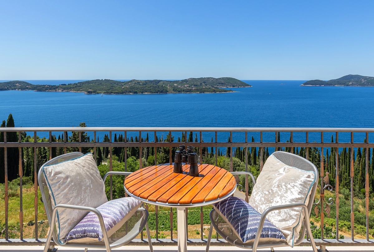 A small wooden table is set with two comfortable chairs on a balcony, offering panoramic views of the sea and surrounding islands. The tranquil blue water contrasts with lush greenery in the distance, creating a serene outdoor space for relaxation.