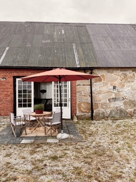 An outdoor seating area features a wooden table surrounded by two chairs, sheltered by a red umbrella. The rustic stone wall and overhanging metal roof provide a charming backdrop. Pebbled ground emphasizes the natural setting, creating a relaxed atmosphere for outdoor enjoyment.
