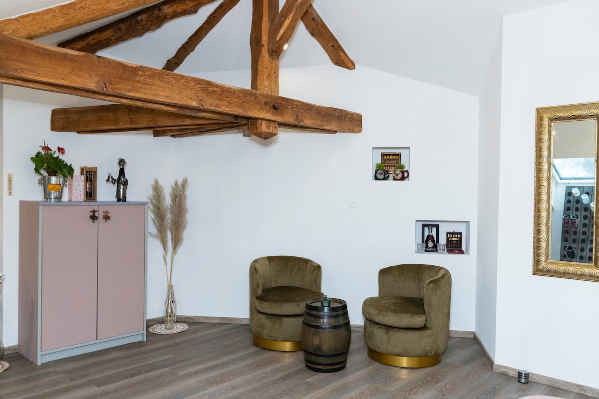 A cozy corner features two olive-green chairs with golden bases, set beside a wooden barrel. A pink cabinet stands nearby, complemented by decorative items on the walls. Natural light fills the space, accentuating the wooden beams of the ceiling.