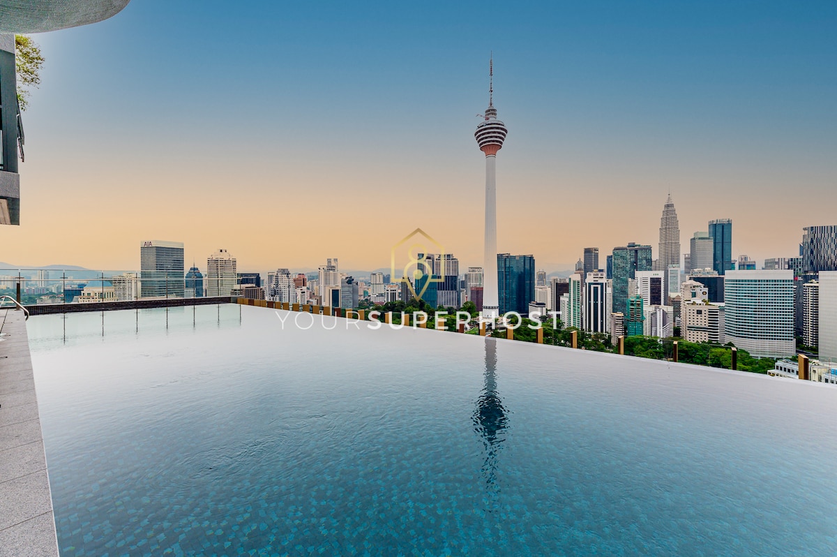 An infinity pool extends towards a breathtaking city skyline, featuring notable buildings including the KL Tower. The water reflects the clear sky, creating a seamless horizon. The vibrant urban landscape is framed by diverse architectural styles, conveying a sense of modernity and tranquility.