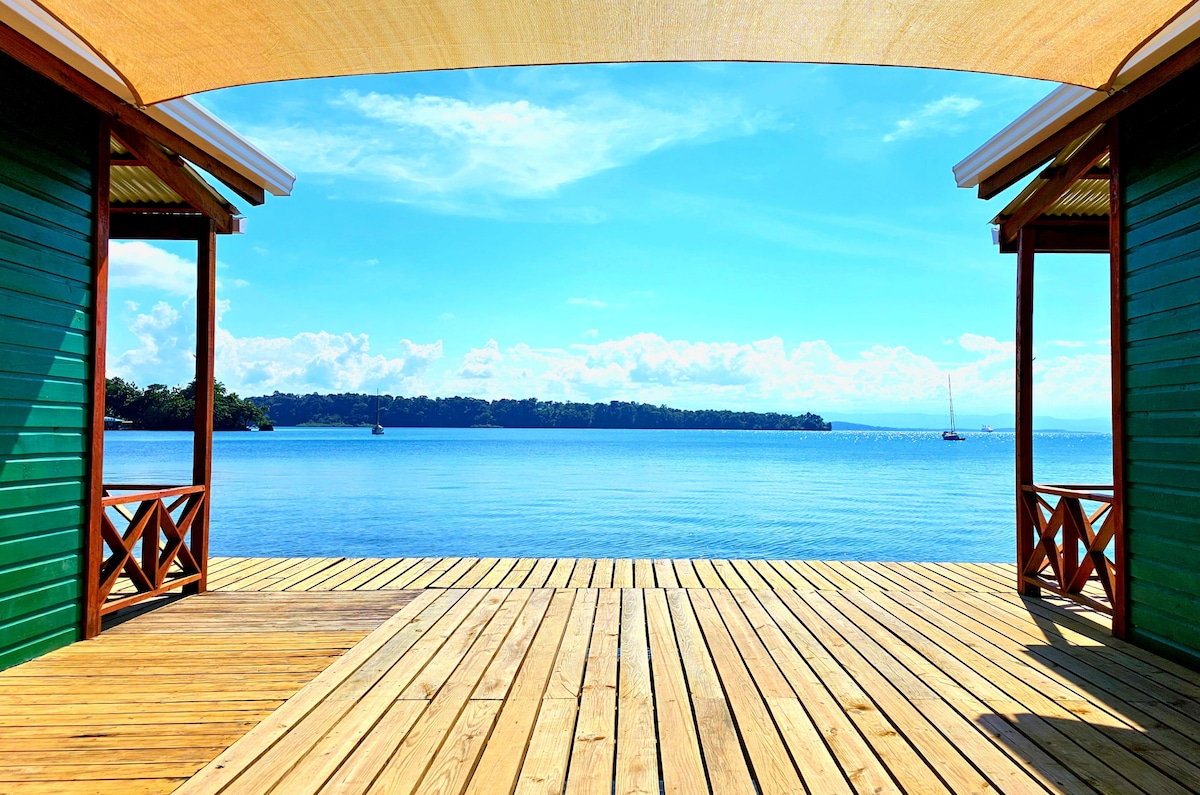 A spacious deck extends over calm waters, framed by two wooden structures. The scene boasts clear blue skies and a panoramic view of the distant shoreline, creating a serene and inviting space for relaxation.