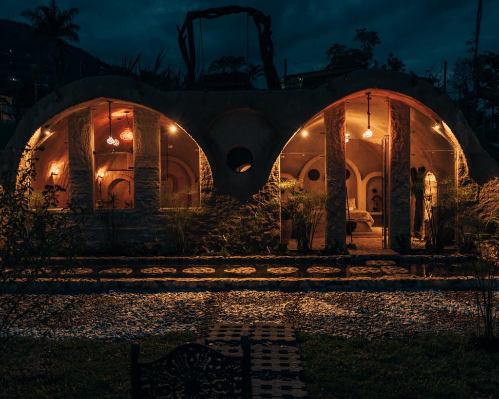 A unique architectural structure is illuminated by soft outdoor lighting, creating a warm ambiance. The building features curved shapes with several archways. Surrounding greenery and a stone pathway provide a natural setting, enhancing the cozy atmosphere of this rural space.