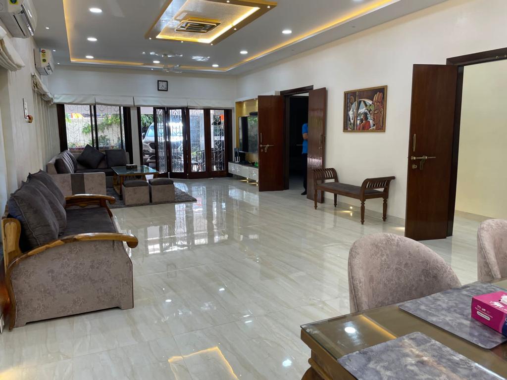 A spacious hall is illuminated with soft lighting, featuring comfortable seating arrangements including plush sofas and chairs. Large glass doors open to an outdoor area, while a dining table is set for six. A television is mounted on the wall, enhancing the communal space.