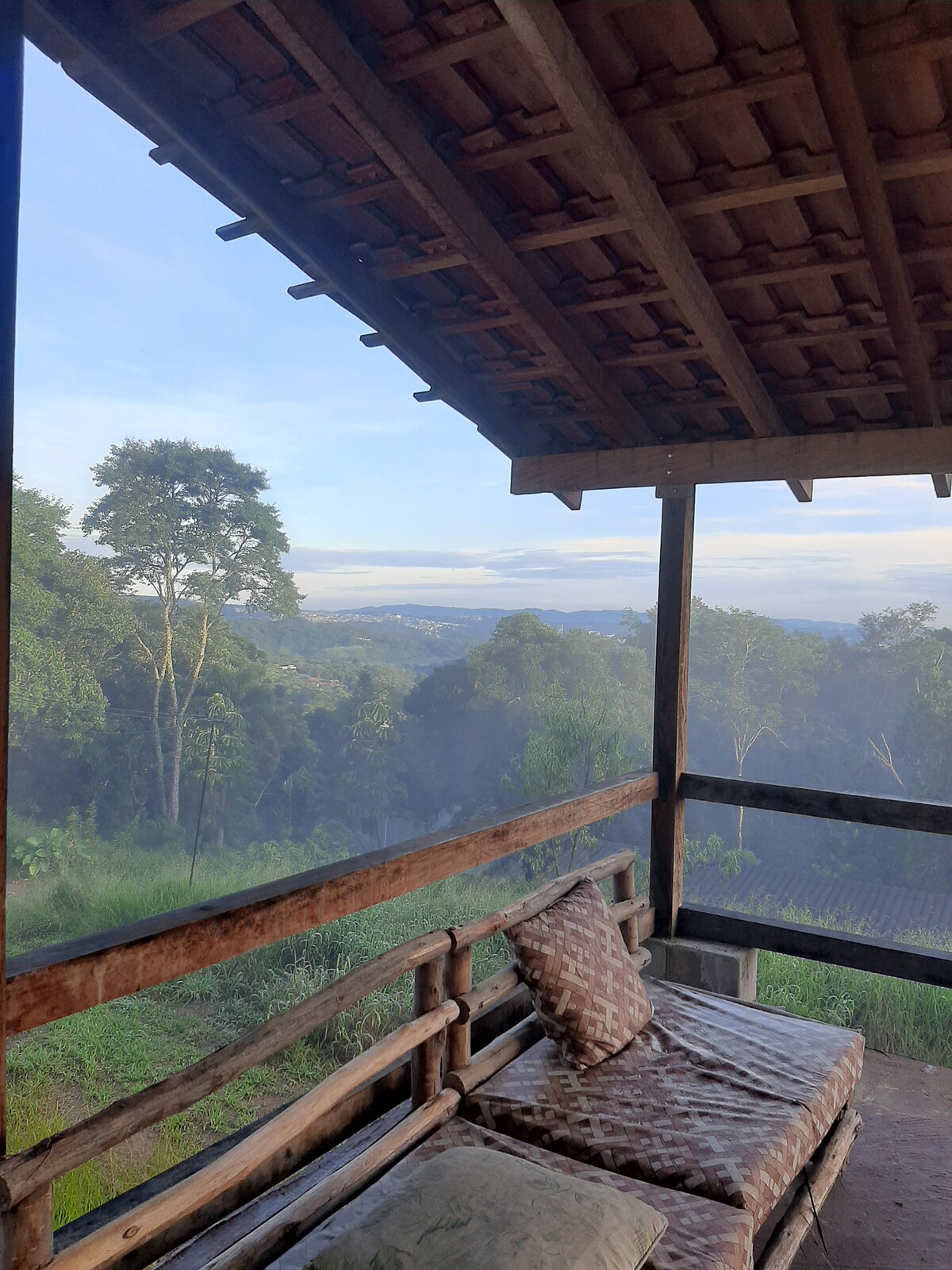 A cozy outdoor seating area is framed by wooden beams, overlooking a misty landscape of lush greenery. Soft cushions adorn the rustic benches, inviting relaxation while enjoying the tranquil view of the rolling hills and trees in the distance.