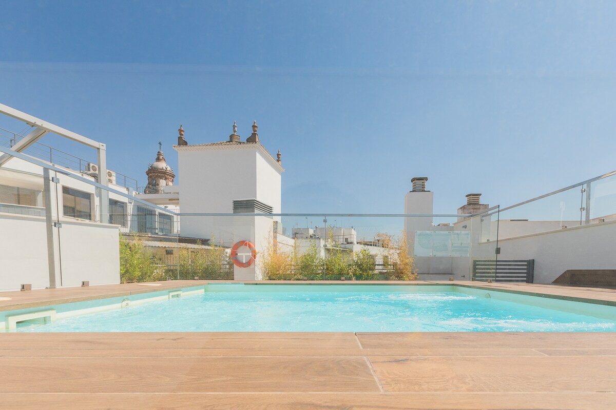 A rooftop swimming pool is depicted, surrounded by wooden decking and featuring clear glass railings. The sunny sky and white rooftops of nearby buildings create an open and bright atmosphere, with a lifebuoy visible for safety.
