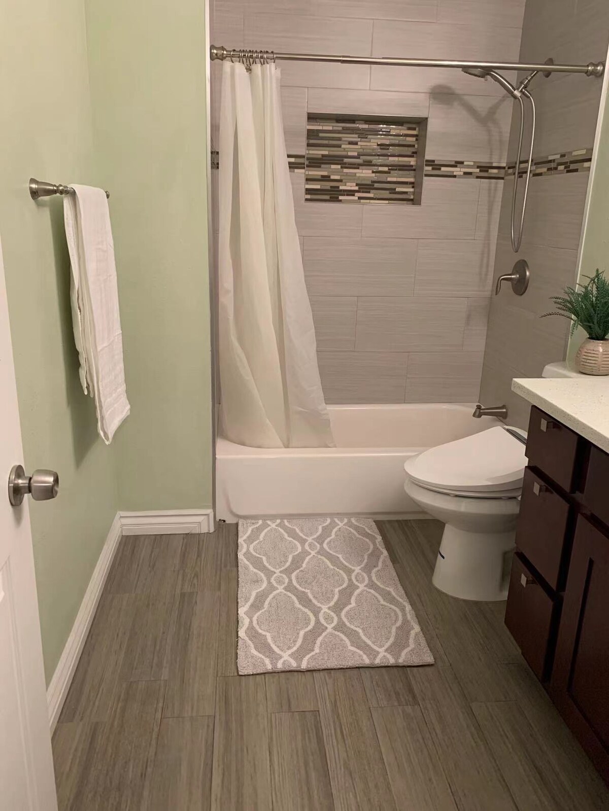 A well-appointed bathroom features a light green wall and a tub-shower combination, with a glass shower curtain. A small rug lies on the floor. A dark wood vanity with a granite countertop is accompanied by a potted plant on top. Fresh white towels are neatly hung.