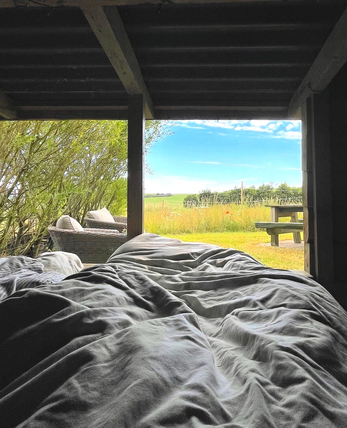 A view from inside the shelter showcases a spacious bed with rumpled bedding. The scene outside features lush greenery and a wooden table. Comfortable seating is visible in the background, framed by an open doorway that invites natural light and fresh air.