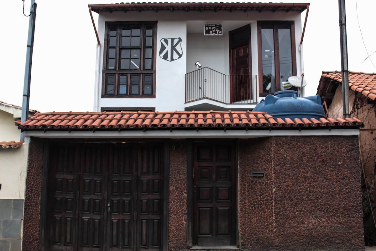 The exterior of the student residence displays a two-story structure with a sloped tile roof. Windows are framed in dark wood, allowing natural light to enter. A balcony with a metal railing is visible on the upper floor, along with satellite dish equipment on the roof.
