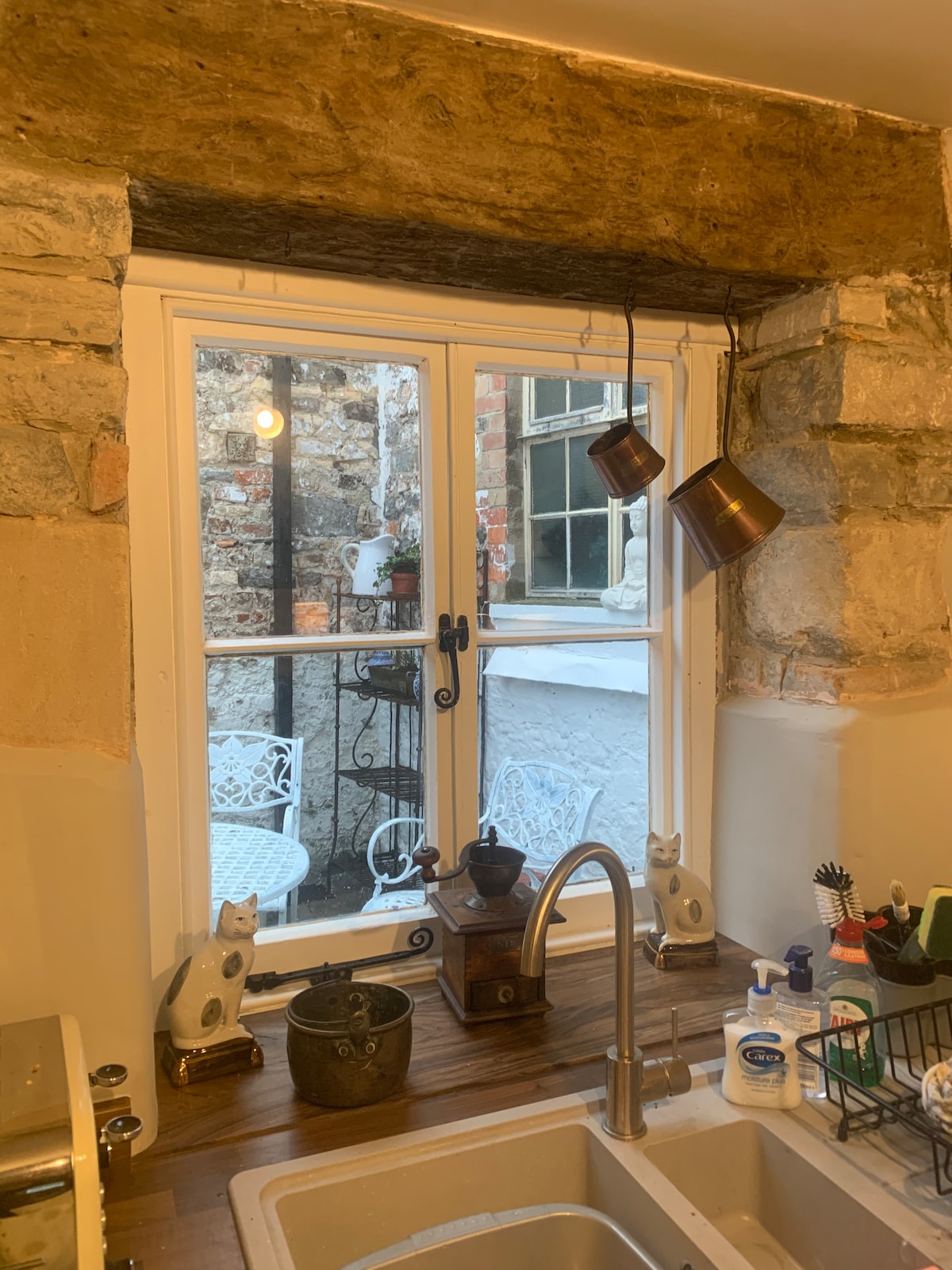 A kitchen window with stone framing offers a view of the peaceful courtyard garden. Two hanging copper pots are visible, alongside charming decorative items on the windowsill. Natural light brightens the kitchen area, highlighting the rustic stone details.