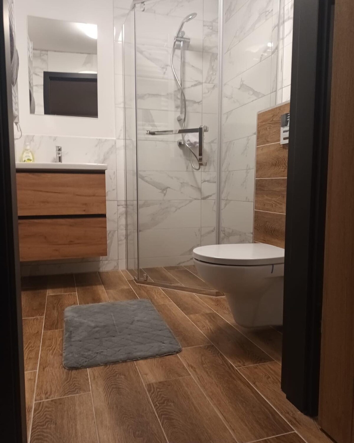 The bathroom features a modern design with wooden cabinetry and a glass-enclosed shower. Light-colored tiles and wooden flooring create a clean and contemporary look. A plush bath mat lies on the floor, and fixtures include a showerhead and a sleek toilet.