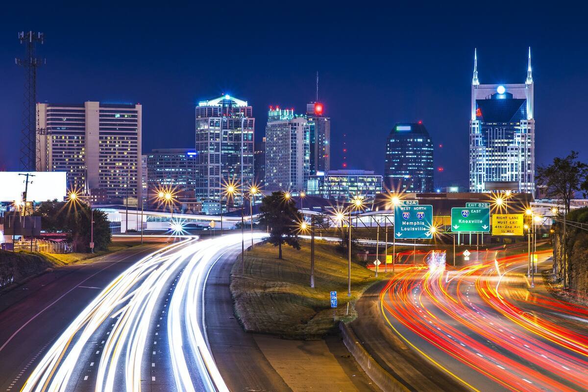 A vibrant city skyline is illuminated against a night sky, featuring numerous tall buildings with twinkling lights. Flowing traffic is visible on the highways below, with streaks of red and white lights indicating movement. Highway signs are positioned prominently, guiding drivers through the urban landscape.