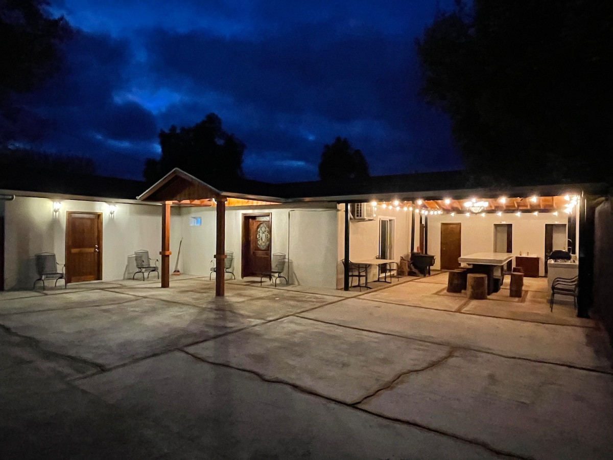 The exterior of the house is illuminated by string lights, creating a cozy ambiance. Concrete flooring spans the area, with several seating options arranged around a central table. The entrance showcases wooden doors, framed by a covered porch, providing access to the main living spaces.