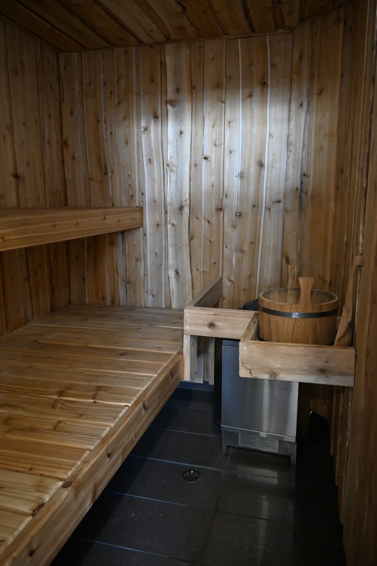 A cedar-lined sauna features a wooden bench along one side and a small stainless steel unit for water heating. A wooden bucket sits nearby, ready for use. The walls are lined with vertical wooden panels, and a stone floor adds a natural feel.