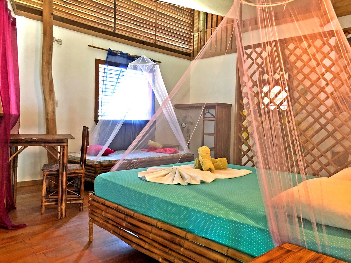 A spacious triple room features a double bed and a single bed, both with mosquito nets. A wooden table and chair are positioned near a window, allowing natural light to fill the space. Brightly colored towels are arranged on the beds, complementing the vibrant decor.