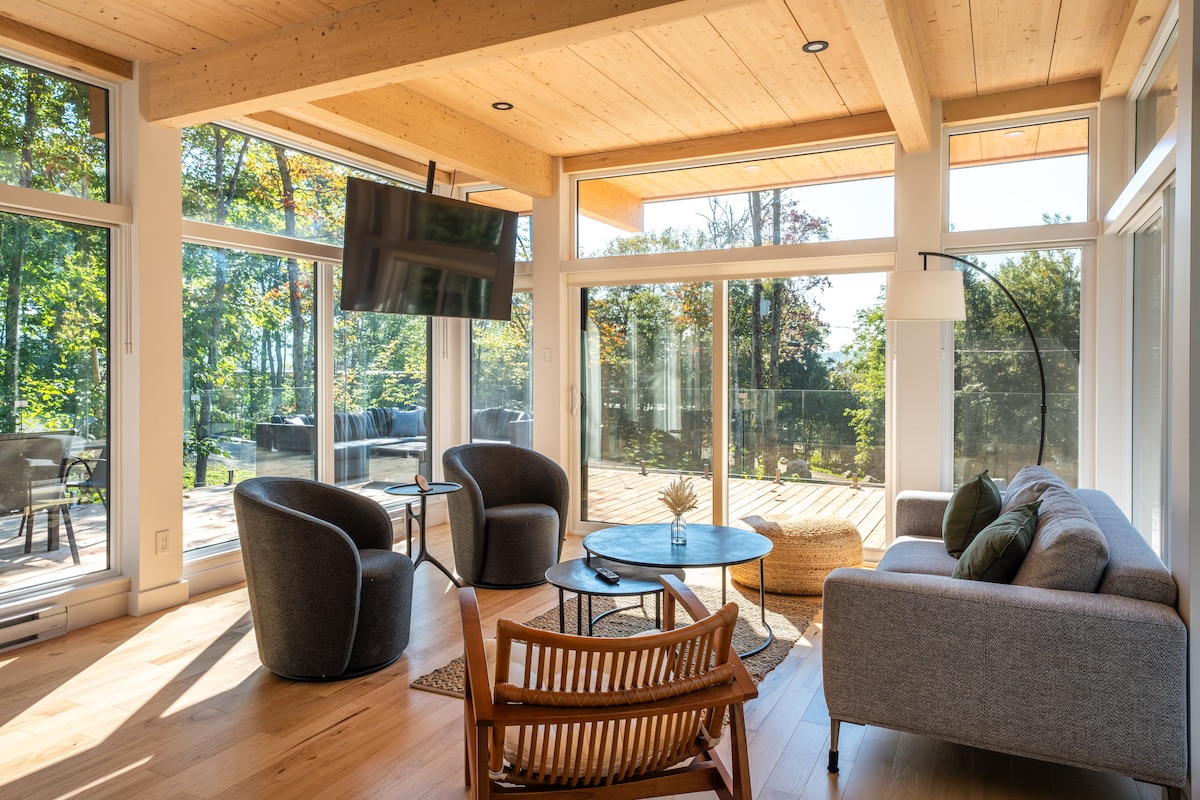 The living area features large floor-to-ceiling windows that provide abundant natural light and views of the surrounding nature. A gray sofa, two accent chairs, and a circular coffee table create a cozy seating arrangement. A wall-mounted TV is visible, enhancing the inviting space.