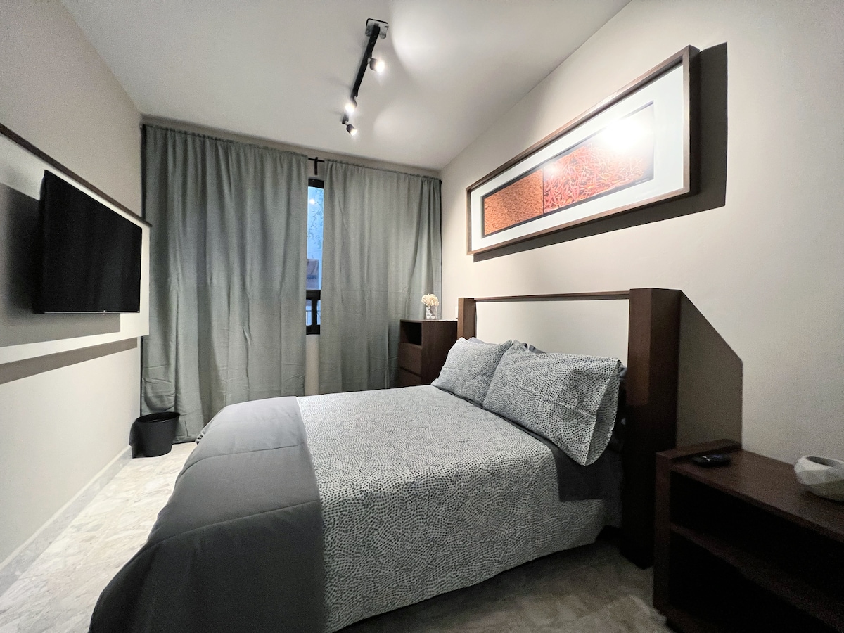 A cozy bedroom is presented with a spacious bed covered in a patterned duvet. Natural light enters through the window, accentuated by soft gray curtains. A wall-mounted television and a bedside table add functionality to the inviting space.
