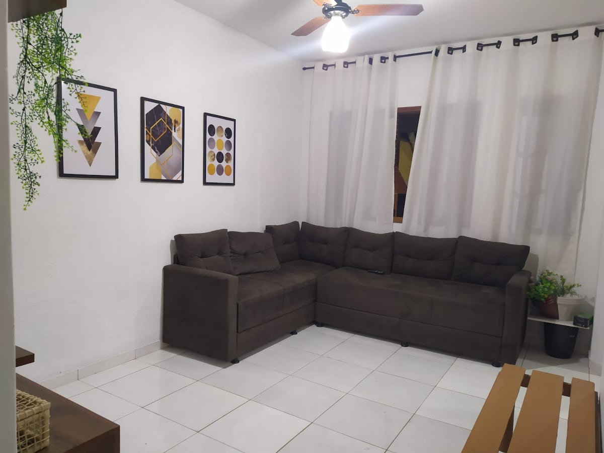 A cozy living area is presented with a spacious sectional sofa in a neutral tone. Three framed wall art pieces add a decorative touch, while a potted plant is positioned nearby. Soft light illuminates the space from a ceiling fan, enhancing the inviting atmosphere.