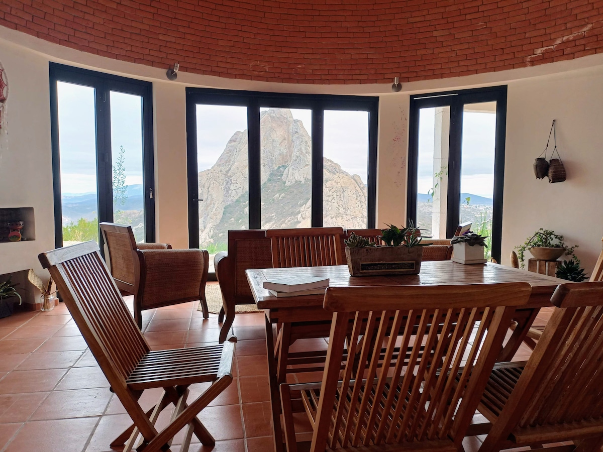 A spacious dining area features a wooden table surrounded by slatted chairs, positioned near large windows that offer views of a prominent mountain. Natural light fills the space, highlighting the warm, earthy tones of the tiled floor and brick walls.