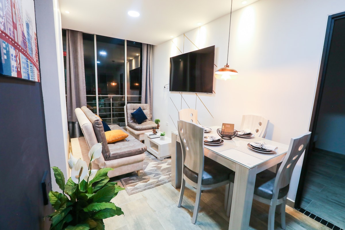 A modern living space is showcased, featuring a comfortable seating area with a gray sofa and bright orange accent pillow. A dining table with four chairs is positioned nearby, complemented by a decorative centerpiece. Large windows allow natural light to fill the room.