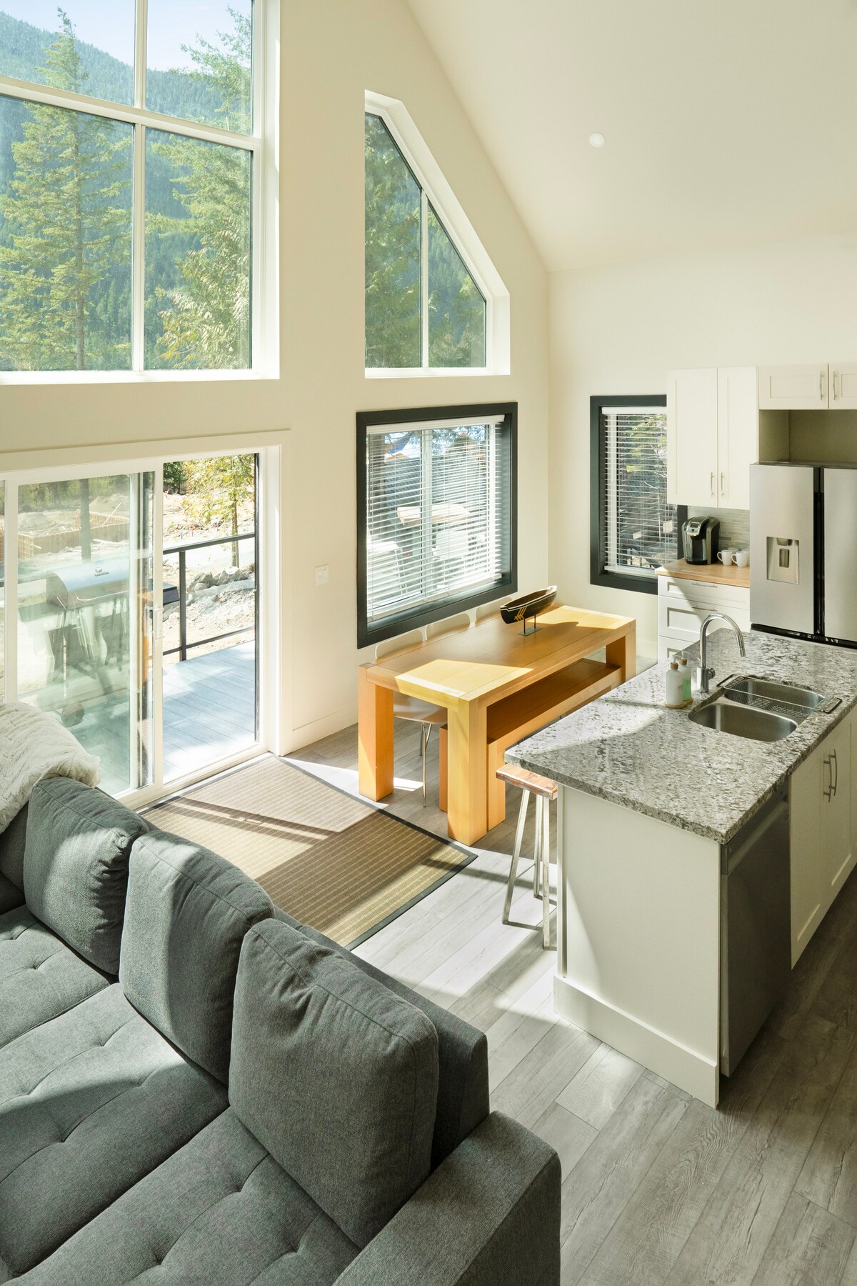 A bright and airy living space features large windows that invite natural light, showcasing views of the outdoors. A cozy sofa is positioned adjacent to a granite countertop kitchen and a light wood dining table is set beside sliding glass doors leading to a balcony.