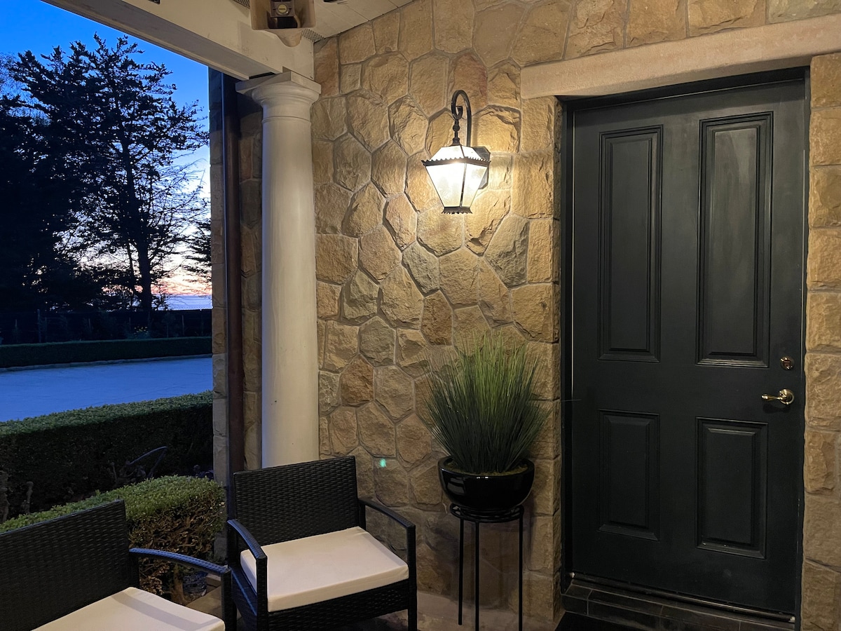 An entryway is featured, adorned with natural stone walls and a decorative lantern. Two black woven chairs with light cushions are positioned on either side of a potted plant. A welcoming dark door presents a contrast to the warm tones of the stonework, with a sunset visible in the background.